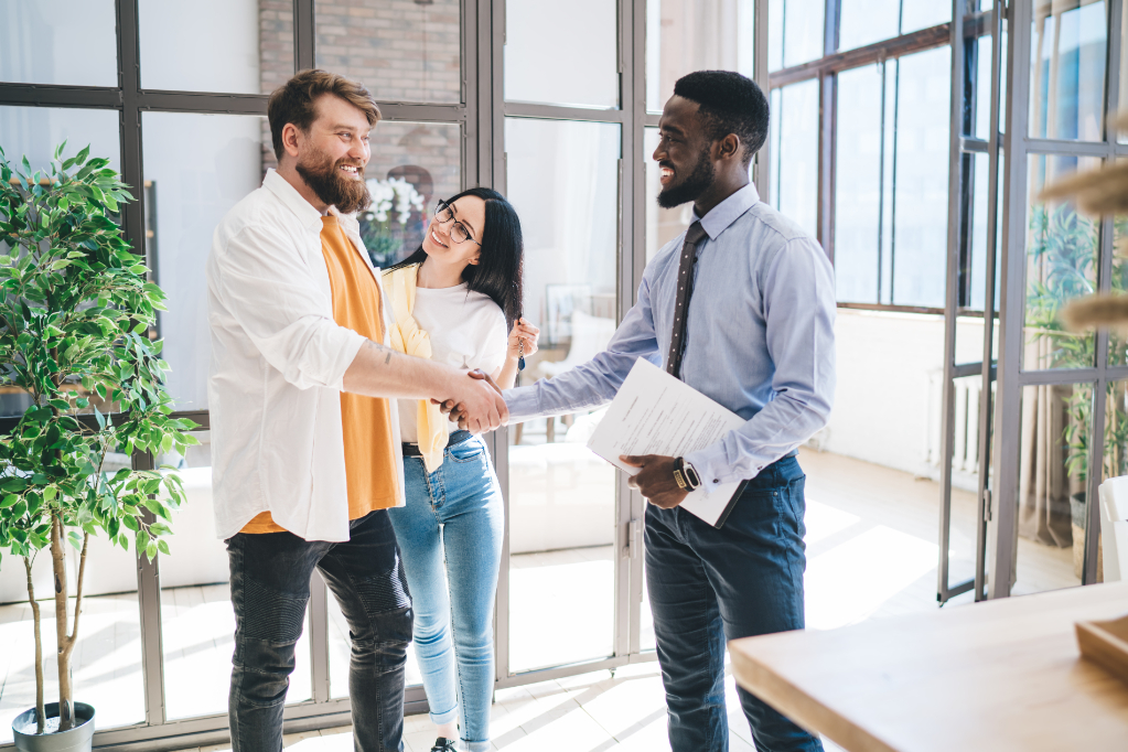 Bearded African American real estate broker with contract and smiling couple sealing deal of apartment purchase by shaking hands while looking at each other